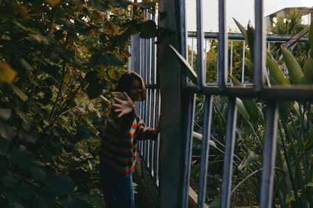 Child girl in a colorful striped sweater reaching hand through a metal gate, curious and smiling, about seven years old, leaning among tropical plants and dense green foliage at dusk.の写真素材