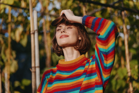 Young woman in her 20s wearing a rainbow sweater and short hair, smiling outdoors in warm sunlight,...