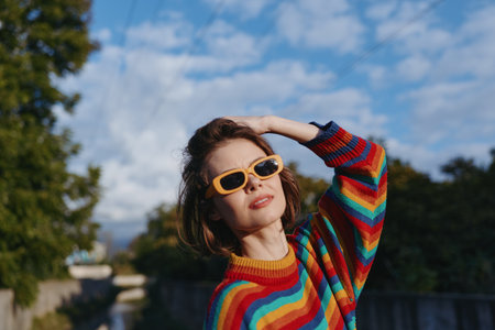 Woman in her 30s with short brown hair wearing yellow sunglasses and a rainbow striped sweater, smiling and posing outdoors by an urban park bridge for lifestyle fashion.の写真素材