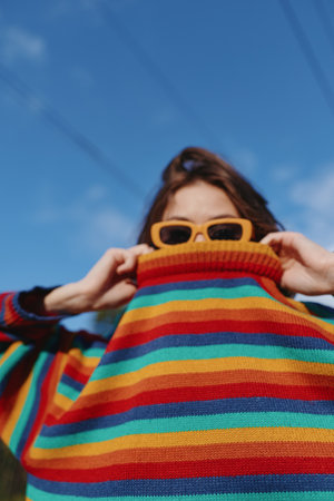 Sweater stripes sunglasses portrait woman colorful fashion against blue sky, playful closeup of person pulling turtleneck over face in outdoor casual style and bright knitwear mood.の写真素材