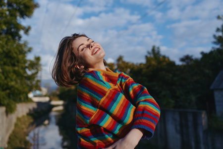 Woman smiling in a rainbow sweater outdoors by a canal, short hair, early twenties, leaning back and laughing, casual outfit, joyful lifestyle portrait in natural lightの写真素材