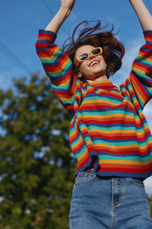 Woman in colorful striped sweater wearing sunglasses smile as she is jumping outdoors under blue sky. Casual jeans, joyful expression, motion and sunny lifestyle portrait.の写真素材