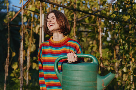 Woman with watering can in garden, smile bright while wearing a colorful striped sweater near greenhouse and vines. Young gardener enjoying sunlight, planting, potting and outdoor hobby activity.の写真素材