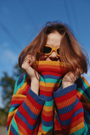 Woman in her twenties wearing a colorful striped sweater and yellow sunglasses, smiling and pulling the knit over her face in a playful outdoor portrait for lifestyle and fashion.の写真素材