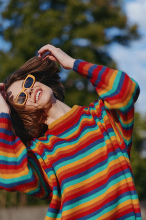 Young woman smiling in sunglasses and a striped sweater outdoors, joyful mid twenties model adjusting retro glasses in sunlit green park, casual outfit and colorful fashion portrait.の写真素材