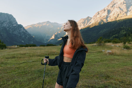 A young woman stands in a mountain valley, rugged jacket and trekking pole in hand, gazing upward....