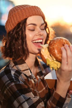 Woman burger smile outdoors beanie plaid enjoying a juicy burger in warm golden hour glow, candid lifestyle portrait with authenticity and emotional storytelling, cozy urban mindful living moment.の写真素材