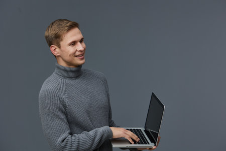 Smiling young man with blonde hair wearing a gray turtleneck sweater holding a laptop isolated on plain gray background. Studio portrait of cheerful professional male. People lifestyle concept.の写真素材