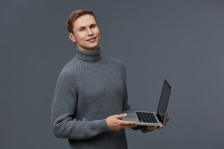 Smiling man holding a laptop in hands wearing a gray knitted turtleneck sweater isolated on plain gray background studio portrait. People lifestyle conceptの写真素材