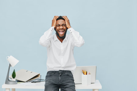Joyful excited young man reacting with surprise and happiness while standing at his modern home office desk, holding his head with both hands, wearing casual white shirt and jeans, cheerful facial expression, isolated on light blue background. People lifestyle conceptの写真素材