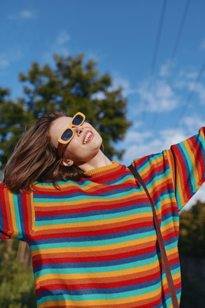 Woman in a rainbow sweater wearing yellow sunglasses, smiling and raising arms outdoors. Young adult casual outfit, joyful expression, leisure lifestyle portrait in bright sunny park.の写真素材