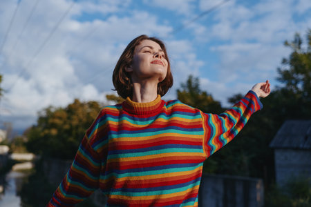 Young woman, short hair, striped sweater smiling with eyes closed outdoors, arms raised enjoying...