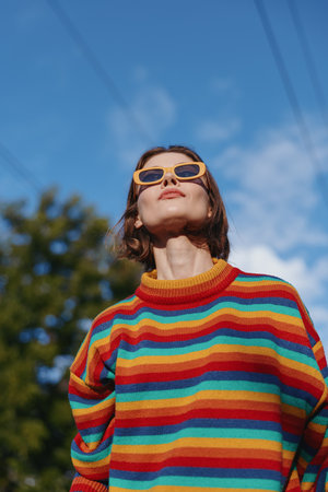 Young adult woman in a colorful striped sweater and yellow sunglasses looking up outdoors against a blue sky, casual outfit, confident pose, lifestyle and fashion portrait.の写真素材