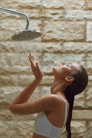 young woman enjoying water shower outdoors with relaxed expression, wet hair, wearing white top, natural stone wall background, refreshing momentの写真素材