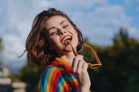 Young woman smiling in a colorful striped sweater, short brown hair, holding sunglasses near her mouth in an outdoor portrait. Casual outfit, joyful lifestyle and sunny mood.の写真素材