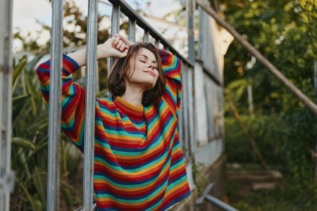 Woman in rainbow sweater smiling and relaxed outdoors, portrait leaning on metal railing among greenery. Casual fashion, cozy knit and warm natural light capturing a peaceful moment.の写真素材