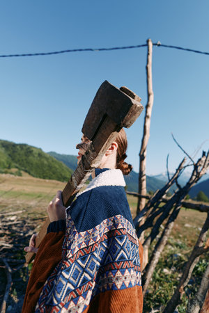 Person carrying an axe on shoulder in patterned jacket in countryside near wooden fence and meadow, mountains in background. Outdoors rural portrait with rustic tool and solitude.の写真素材