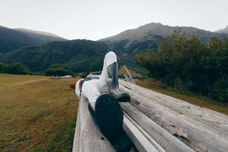 Person mountains meadow wooden relax outdoor lying on a rustic log bench in alpine field, traveler resting with boots and jacket, peaceful nature escape and wide scenic view.の写真素材