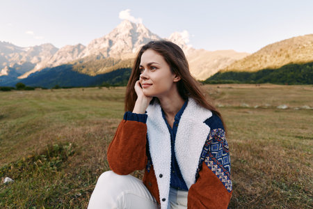 Woman portrait sitting in meadow with mountains in background, smiling and relaxed in a cozy jacket....