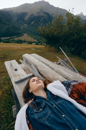 Woman lying on a wooden bench in the mountains nature, relax outdoors in a meadow wearing cozy jacket and denim shirt, portrait of peaceful rural escape and scenic landscapeの写真素材