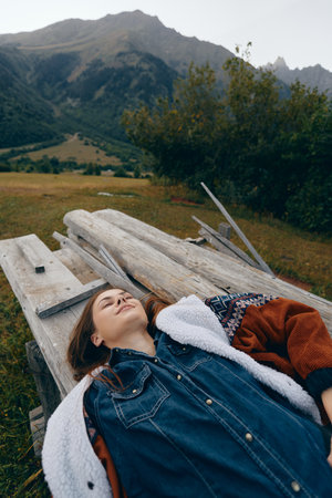 Woman lying on wooden bench in nature near mountains, portrait of relaxation in meadow with jacket and closed eyes, outdoors escape, calm scenery and peaceful rural landscape.の写真素材