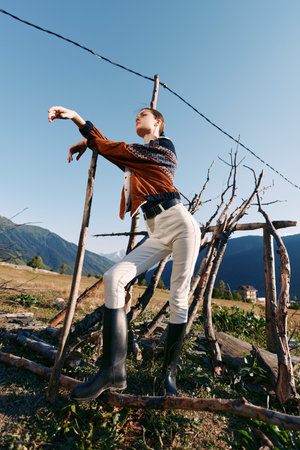 Woman model posing by rustic fence in countryside wearing rubber boots and patterned sweater, stylish outdoor fashion portrait with mountains background and confident stanceの写真素材