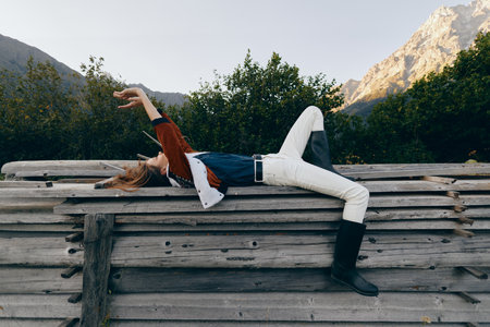 Woman lying on wooden fence relaxing outdoors with mountains in background, casual outfit and boots....
