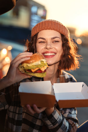 Woman holding a burger outdoors, smile and candid takeout lifestyle portrait in goldenhour with cozy beanie and plaid, authenticity, emotional storytelling and mindful living capture the moment.の写真素材