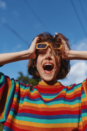 Woman portrait in a colorful sweater with sunglasses shouting joyfully outdoors, fashion look and energetic expression, rainbow stripes, short hair and sunny blue sky backdrop.の写真素材