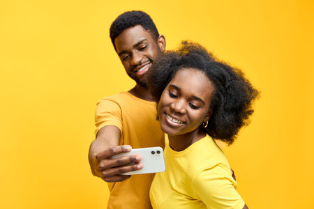 Young cheerful African couple taking a selfie with a smartphone in vibrant yellow shirts against a bright yellow background, capturing joyful emotionsの写真素材