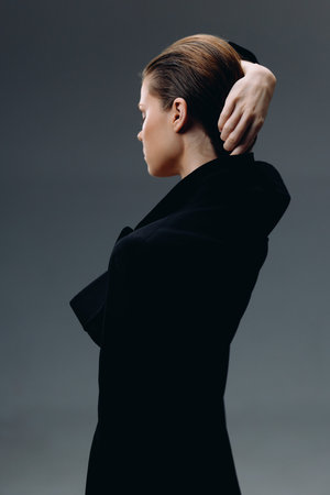 Elegant woman in black coat posing with hand behind head against gray backgroundの写真素材