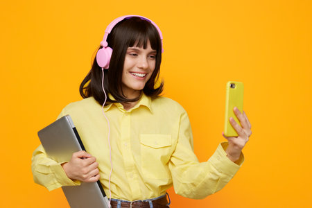 A cheerful young woman wearing pink headphones and a yellow shirt, holding a laptop and smartphone while posing against a bright orange background.の写真素材