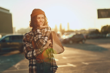 Woman shopping with smile at sunset in urban parking, candid portrait of beanie-wearing model holding shoppingbag. Authenticity, candid lifestyle, golden hour glow, mindful living.の写真素材