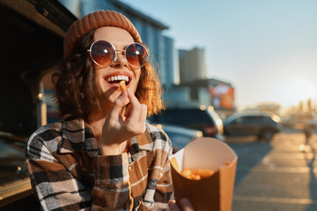 woman eating fries from takeout by car trunk at sunset, sunglasses and beanie, genuine smile in a candid lifestyle scene with golden hour glow, authenticity, mindful living and emotional storytellingの写真素材