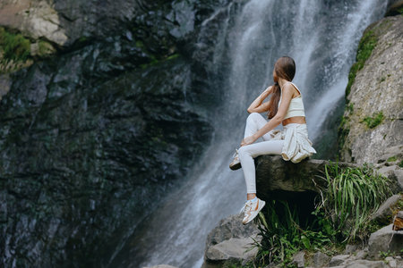 Young woman sitting on rock near waterfall, wearing white casual clothes and sneakers, enjoying nature and peaceful outdoor environment during daytime.の写真素材
