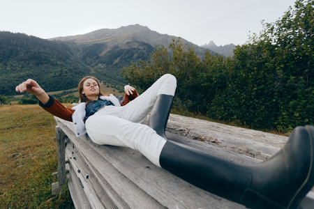 Woman in rubber boots relaxing on a wooden bench in a meadow with mountain backdrop, casual outfit and peaceful nature scene for outdoor leisure, hiking rest and countryside escapeの写真素材