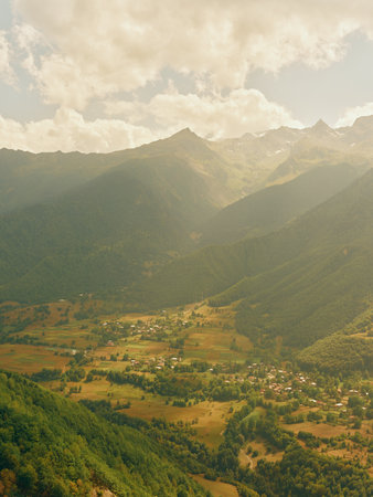 Mountain valley village landscape with rugged peaks and soft clouds, aerial countryside view showing green forest, meadows and scattered houses in warm golden sunlight.の写真素材