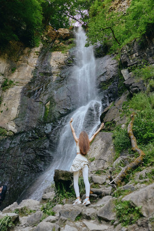 woman enjoying waterfall in forest surrounded by lush greenery and rocky terrain with sunlight filtering through trees for outdoor adventureの写真素材