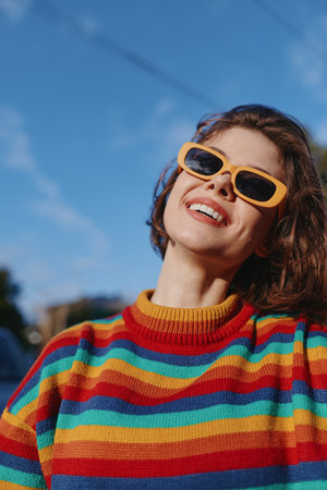 Woman smiling in a colorful striped sweater and yellow sunglasses, outdoor sunny portrait of a mid adult casual model leaning back with joyful expression for travel lifestyle fashion use.の写真素材