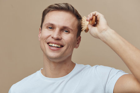 Smiling man using a brush to groom his hair, casual light blue t-shirt, cheerful expression, indoor background, lifestyle moment, male grooming, everyday routine, positive mood, natural lighting, portrait shot.の写真素材