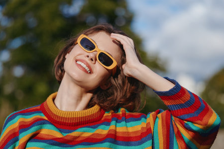 Woman smiling in a colorful striped sweater and yellow sunglasses, short hair, outdoor portrait; casual outfit, hand in hair, joyful expression, lifestyle fashion travel vibe.の写真素材