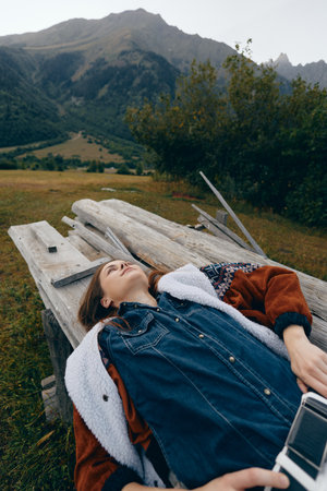 Woman lying on a wooden bench in a mountain meadow, relaxed and contemplative in a denim jacket holding a camera. Scenic landscape and peaceful outdoor retreat with distant peaks.の写真素材