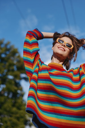 Woman rainbow sweater sunglasses smiling outdoors portrait of joyful young person under blue sky, colorful knit fashion, casual jumper and trendy eyewear conveying carefree summer mood.の写真素材