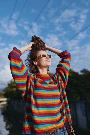Young woman in her 20s wearing a colorful striped sweater and yellow sunglasses, smiling outdoors...