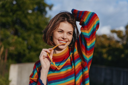 Woman with a bright smile, portrait in a colorful sweater outdoors holding sunglasses and posing casually, natural light, short hair, playful cozy autumn fashion and joyful expression.の写真素材