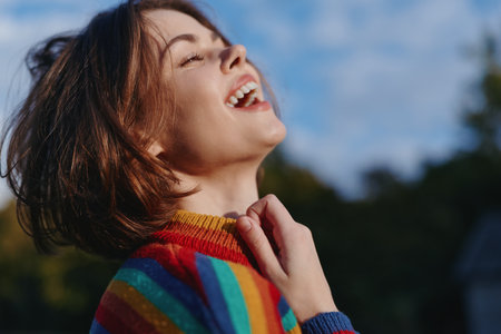 Young adult woman (mid 20s) smiling and laughing in a colorful rainbow sweater, short hair portrait outdoors at golden hour, joyful mood for lifestyle and fashion travel.の写真素材