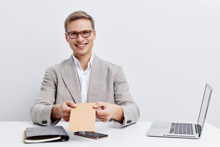 Smiling mature man in glasses and light grey blazer sitting at desk with electronic devices and notepad, holding a piece of paper, studio shot on plain background. Professional, business, office, productivity concept.の写真素材