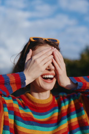 woman smiling striped sweater sunglasses covering eyes outdoor young adult joyful: Young adult woman smiling in a colorful striped sweater with sunglasses on head, covering eyes with hands whileの写真素材
