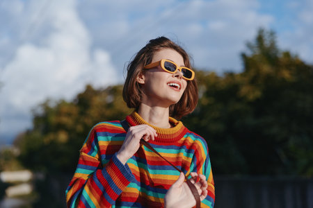 Woman smiling in a colorful striped sweater and yellow sunglasses, young woman enjoying outdoor portrait in park, short bob hair, casual outfit, looking up, joyful travel lifestyle and fashion moment.の写真素材