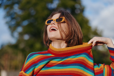 Woman smiling in a colorful striped sweater and yellow sunglasses, casual short hair portrait outdoors, joyful young adult pulling collar, lifestyle and fashion travel vibe.の写真素材